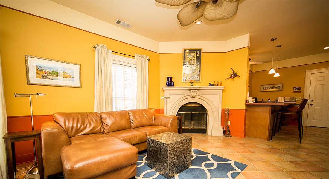 Spacious and open living room off of kitchen decorated in warm yellow. Fireplace with mantle beneath artwork and wall hangings.