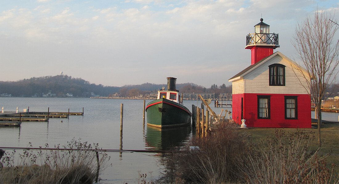 Exterior view of red and white lighthouse next to green and white boat on large expanse of water with hills in the background.