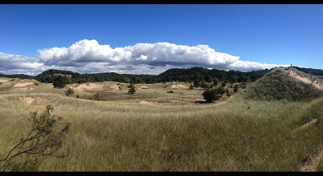 Open view of white thunderclouds forming over distant hills as viewed over rolling plains with praire grass leading into green woods.