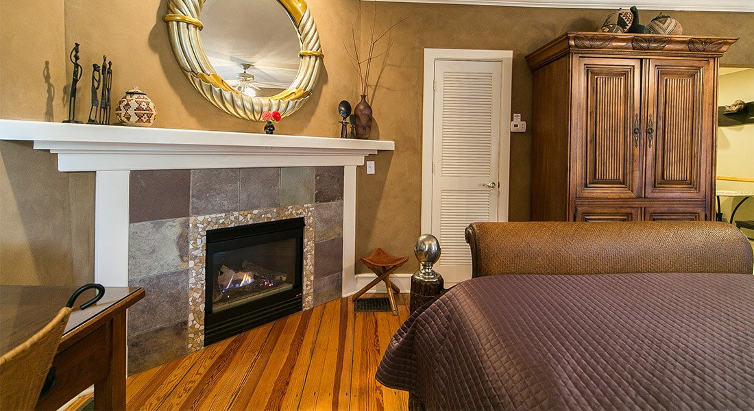 Gold accent mirror hangs above gas fireplace focal point. Armoir and entry to kitchen can be seen over sleigh bed.