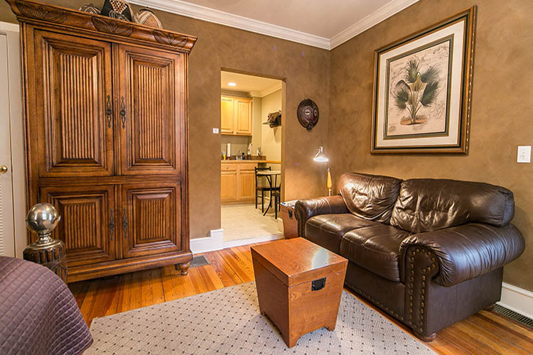 Brown leather couch and box-table underneath framed artwork flanking a door to kitchenette with a multi-level armoire.