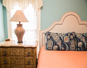 Bed with peach comforter, tufted headboard, and lamp sitting on bedside dresser in front of window.