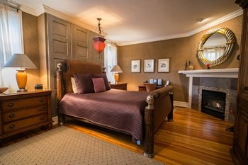 Bedroom with high back bed with purple spread. Wood floors, and fireplace: Link leads to Rooms page