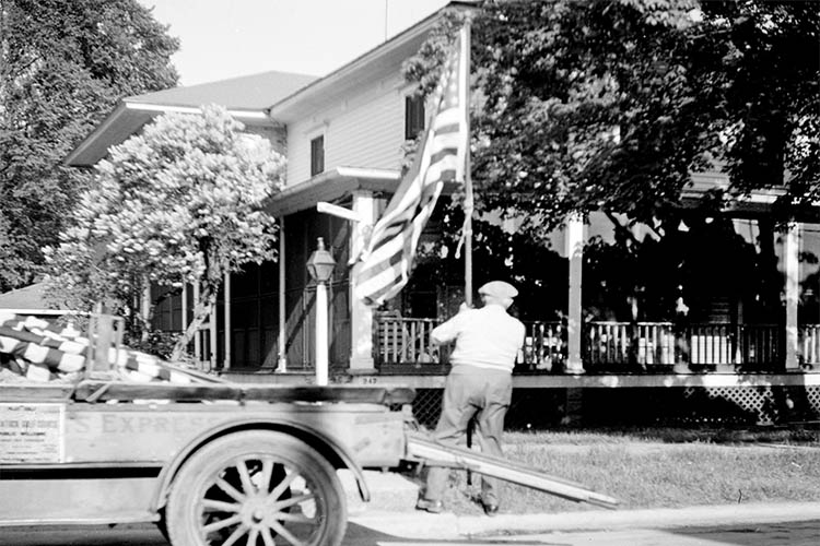 Old picture with man putting up flag in front of old house and car.