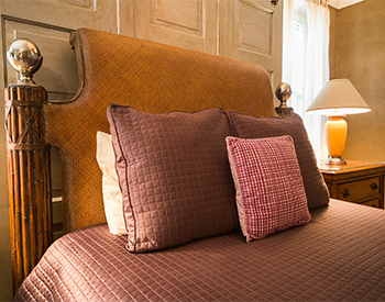 Deep red spread and pillows against a tan head board with amber lamp on bedside table underneath window.