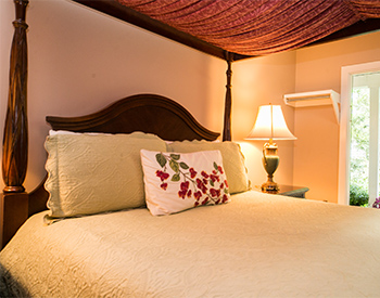 Four-Post canopy bed with burgandy drape hanging over beige bedspread and decorative pillows with lamp on bedside table.