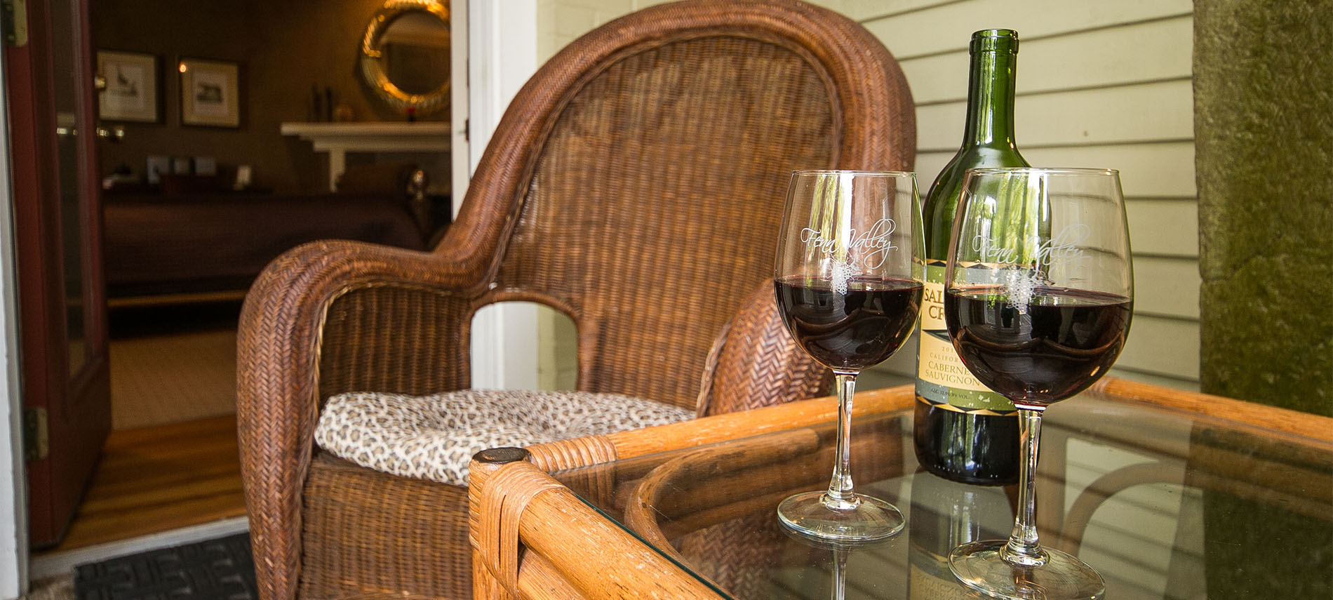 Wine bottle and two glasses of wine sitting next to a wicker chair with door to room open.