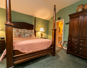 Four-post high pillar bed with pink afghan and floral pattern pillows. Antique armoire stands in the corner.